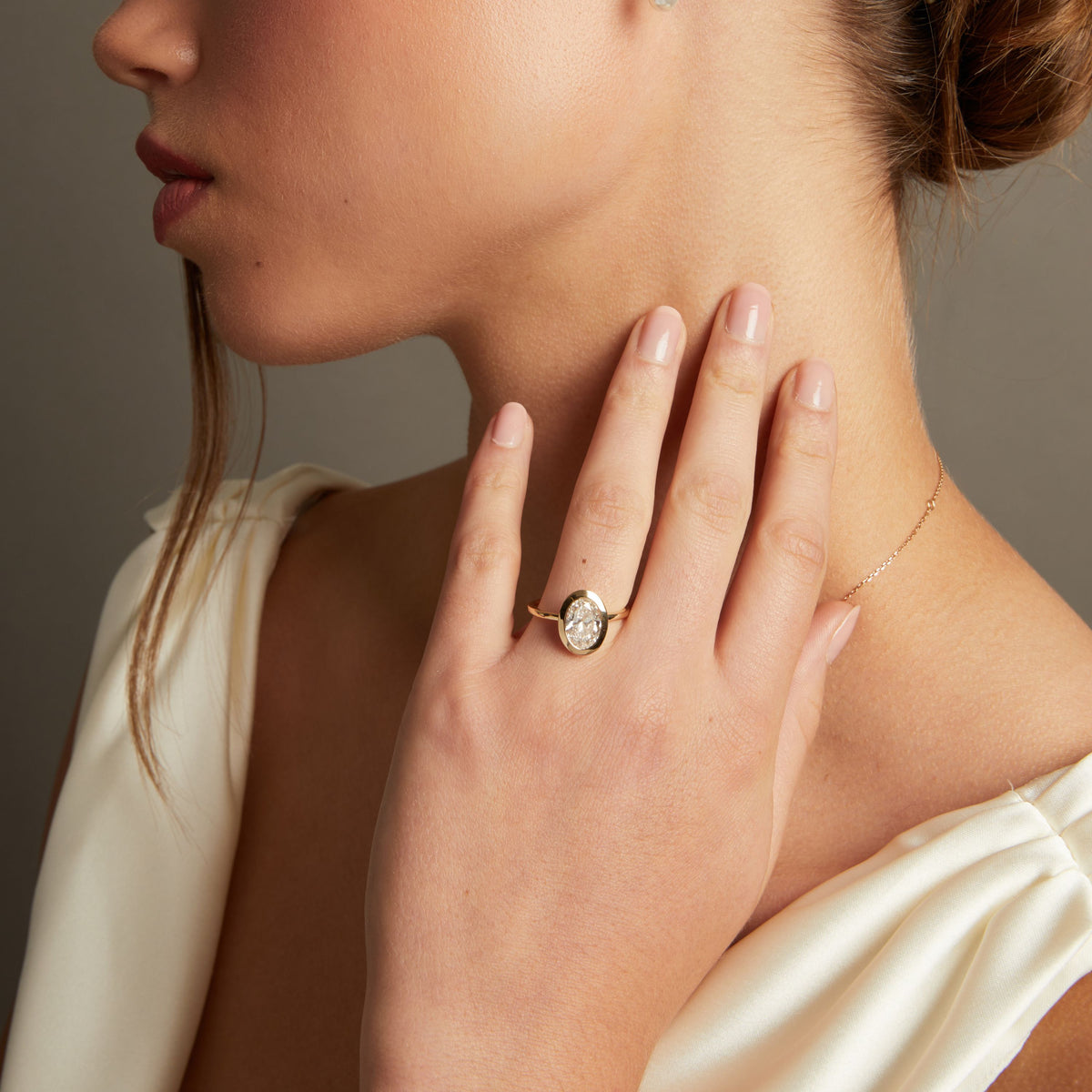 Model. A close-up lifestyle shot of a woman wearing an oval shaped diamond bezel ring on her finger, paired with a delicate gold necklace against a neutral gray background.