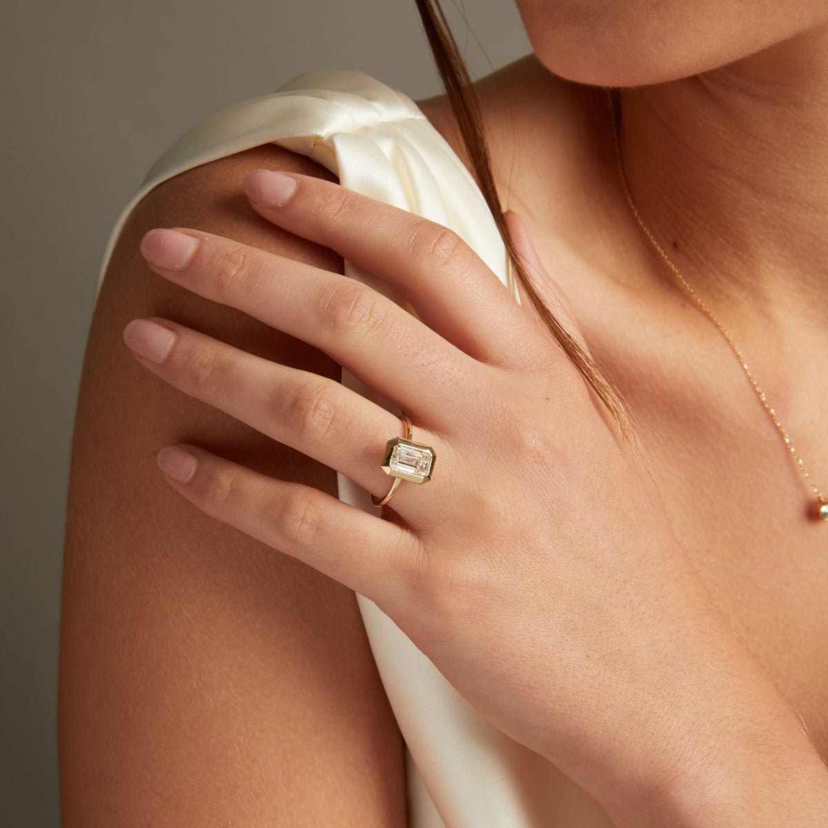 Model. A close-up lifestyle shot of a woman's hand wearing a gold ring with an emerald bezel-set diamond. She is wearing a cream satin top against a neutral gray background.