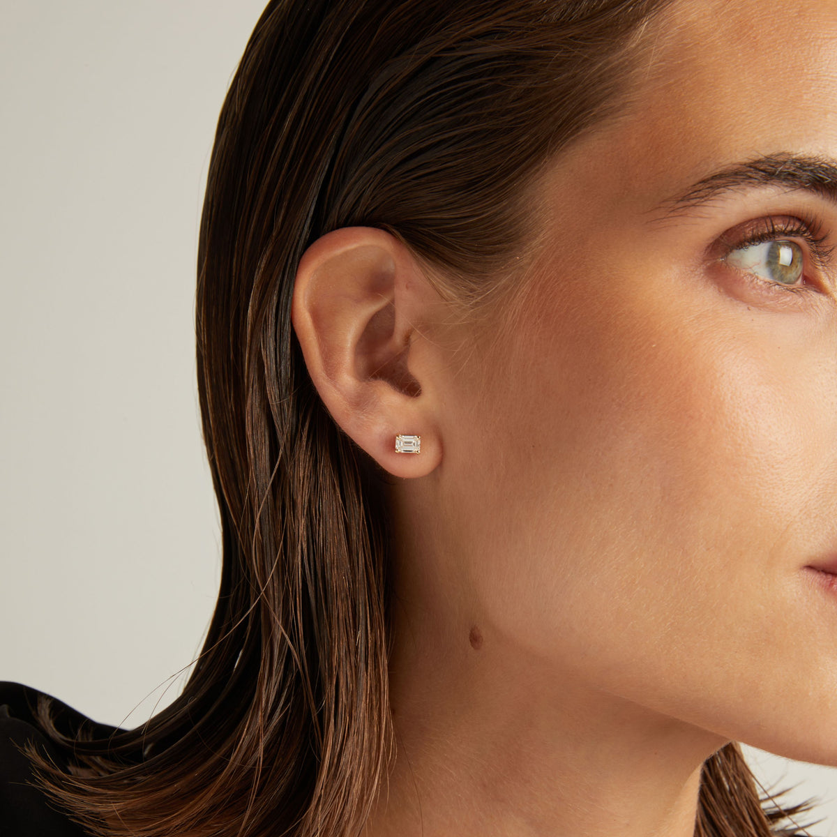 Model. Close up of woman wearing an emerald shaped diamond stud earring on a beige background.