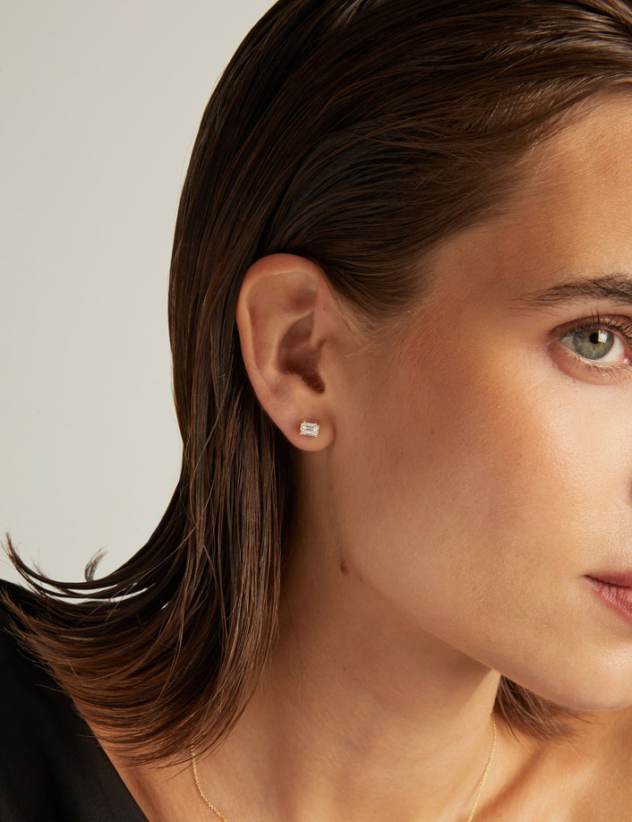 Model. Close up of woman wearing an emerald shaped diamond stud earring on a beige background.