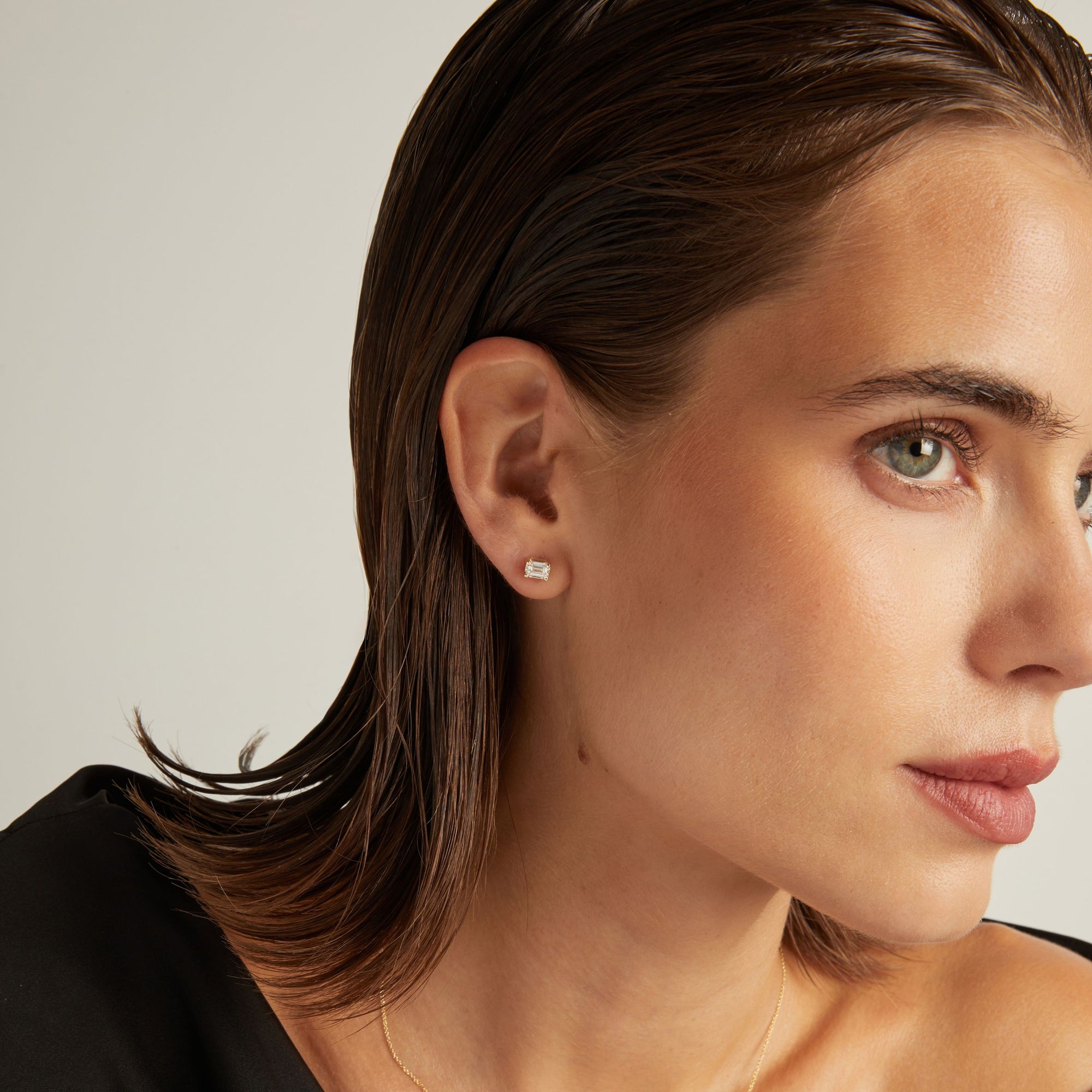 Model. Close up of woman wearing an emerald shaped diamond stud earring on a beige background.