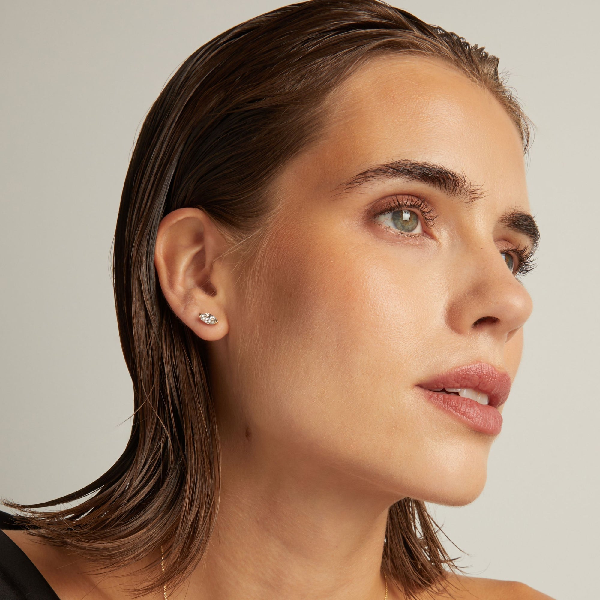 Model. Close up of woman wearing a marquise shaped diamond stud earring on a beige background.