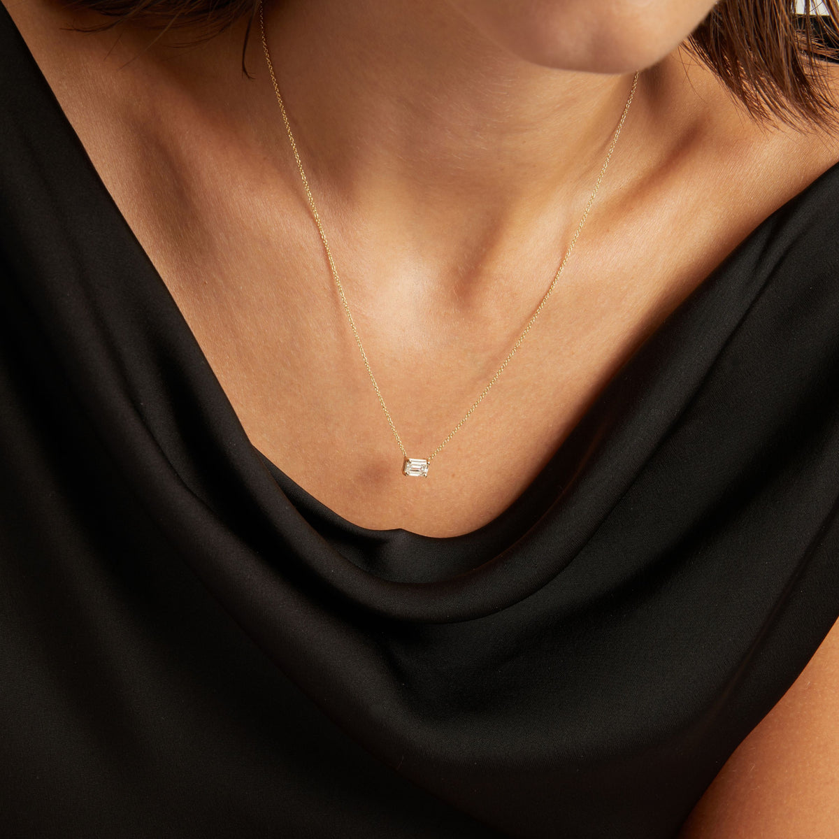 Model. Close up of woman wearing a black top with an emerald shaped diamond solitaire necklaces on a beige background.