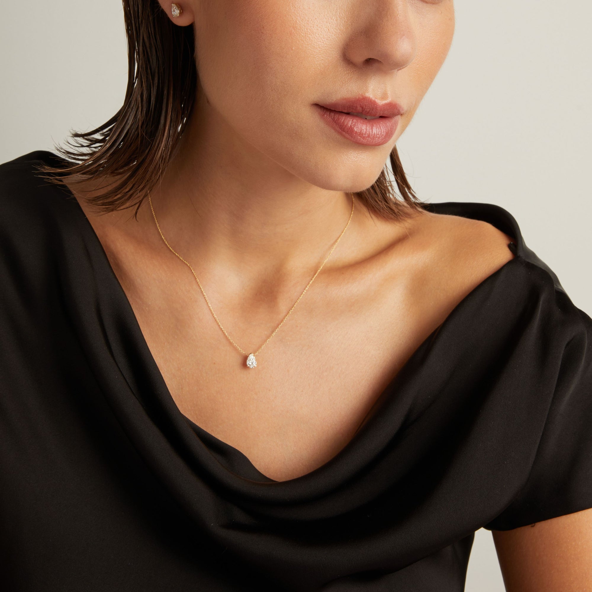Model. Close up of woman wearing a black top with a pear shaped diamond solitaire necklaces on a beige background.