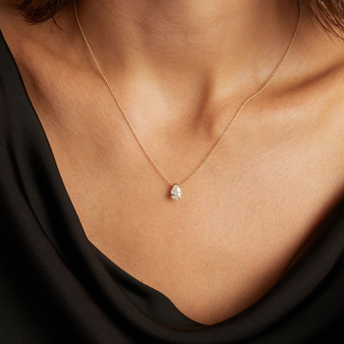 Model. Close up of woman wearing a black top with a pear shaped diamond solitaire necklaces on a beige background.