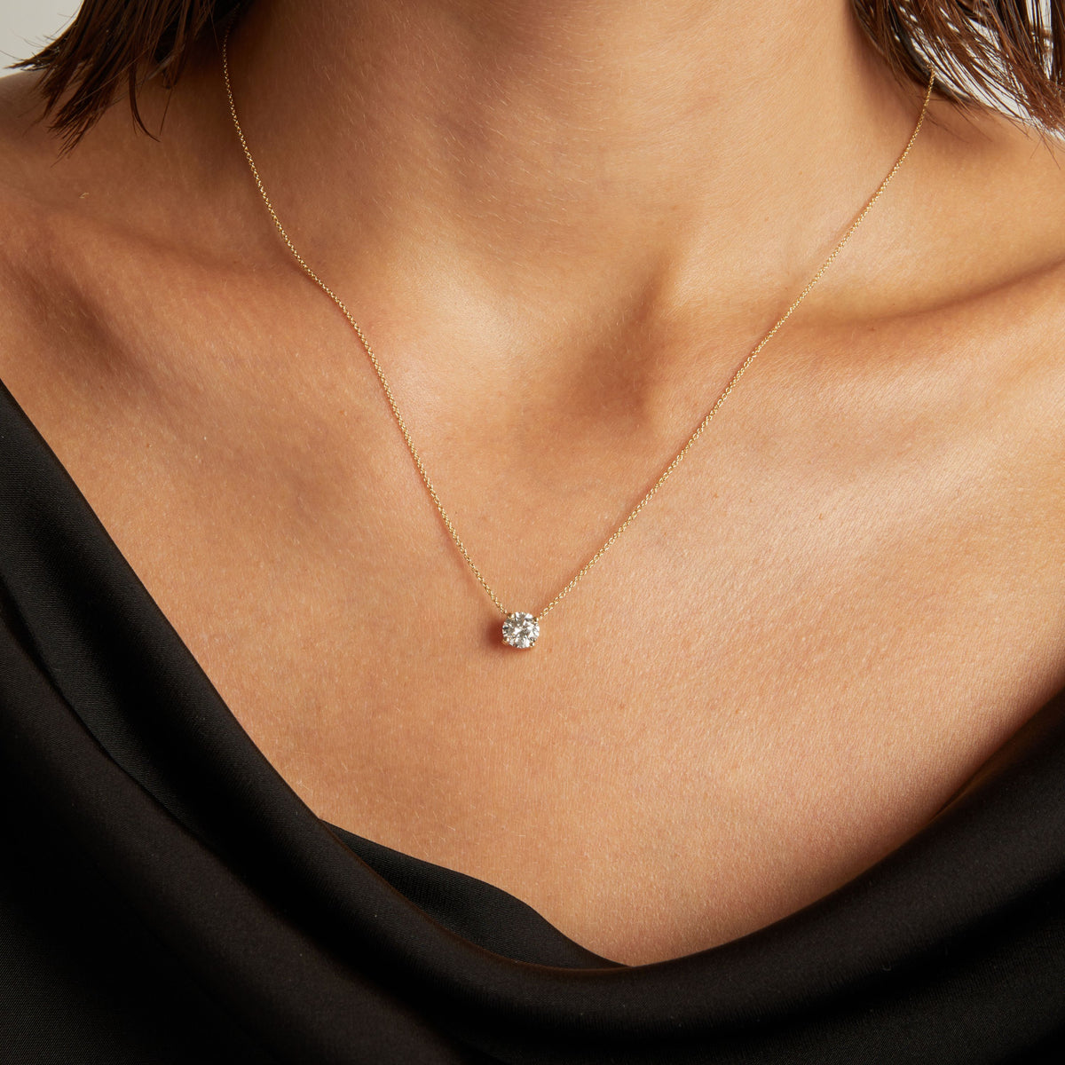 Model. Close up of woman wearing a black top with a round shaped diamond solitaire necklaces on a beige background. 