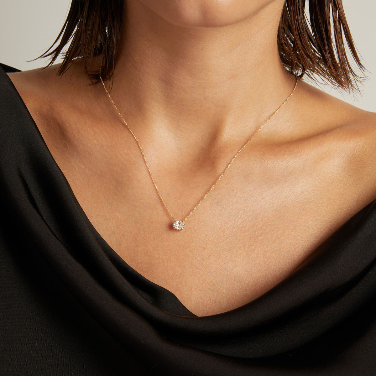 Close up of woman wearing a black top with an oval shaped diamond solitaire necklaces on a beige background.