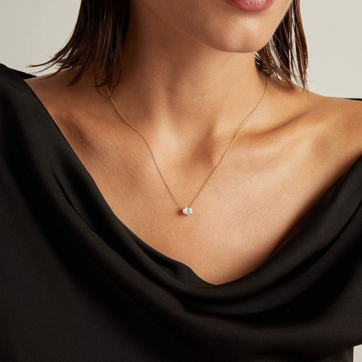 Model. Close up of woman wearing a black top with an oval shaped diamond solitaire necklaces on a beige background.