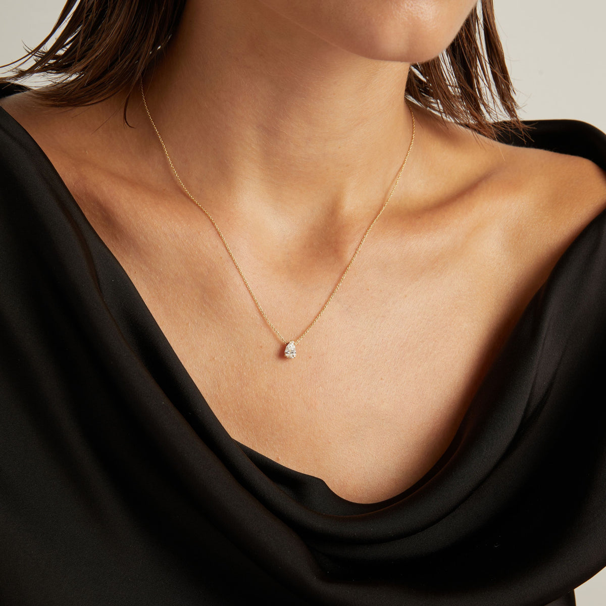 Close up of woman wearing a black top with a pear diamond solitaire necklaces on a beige background.