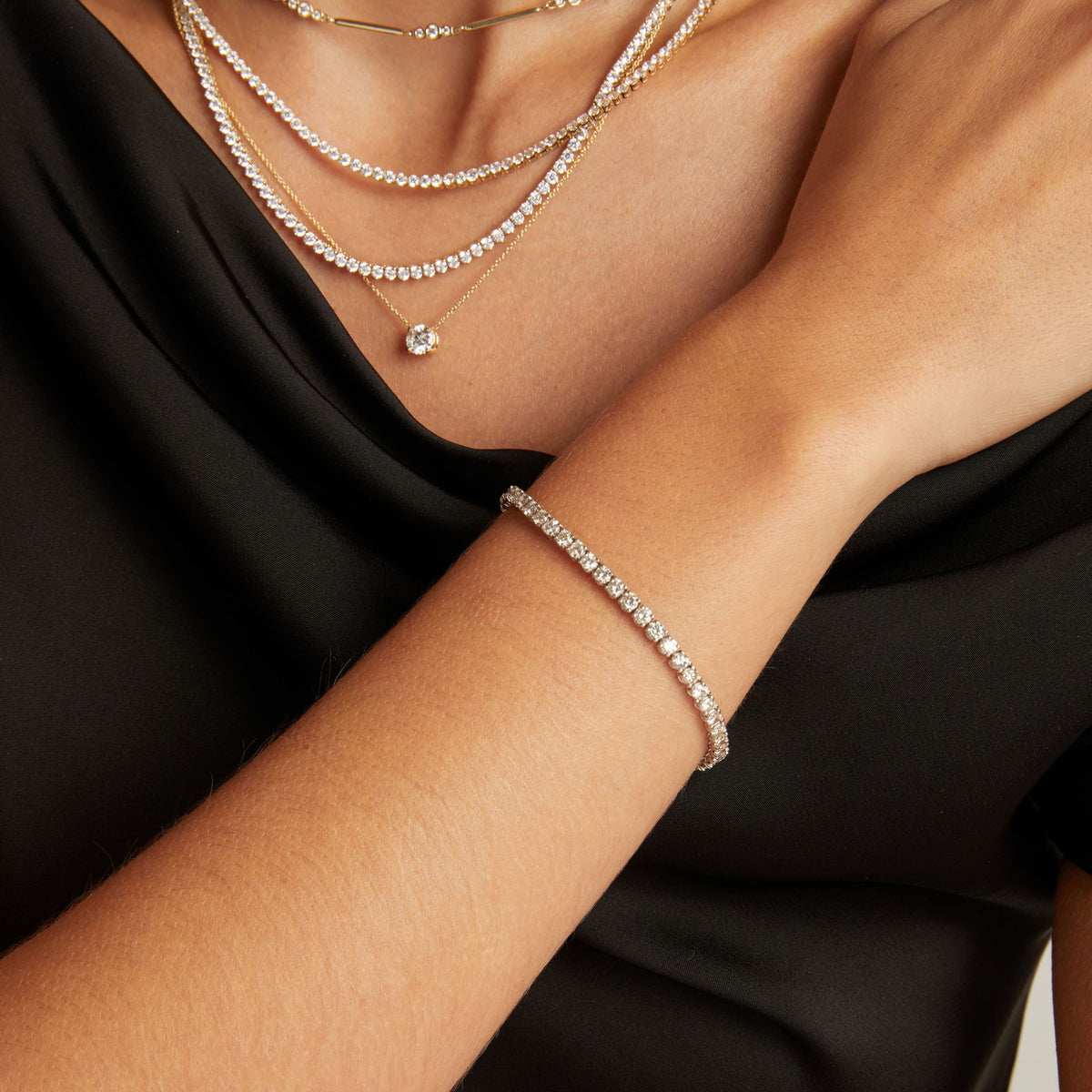 Model. Close up of a woman wearing a black top with stacked diamond necklaces and a diamond tennis bracelet on beige background.