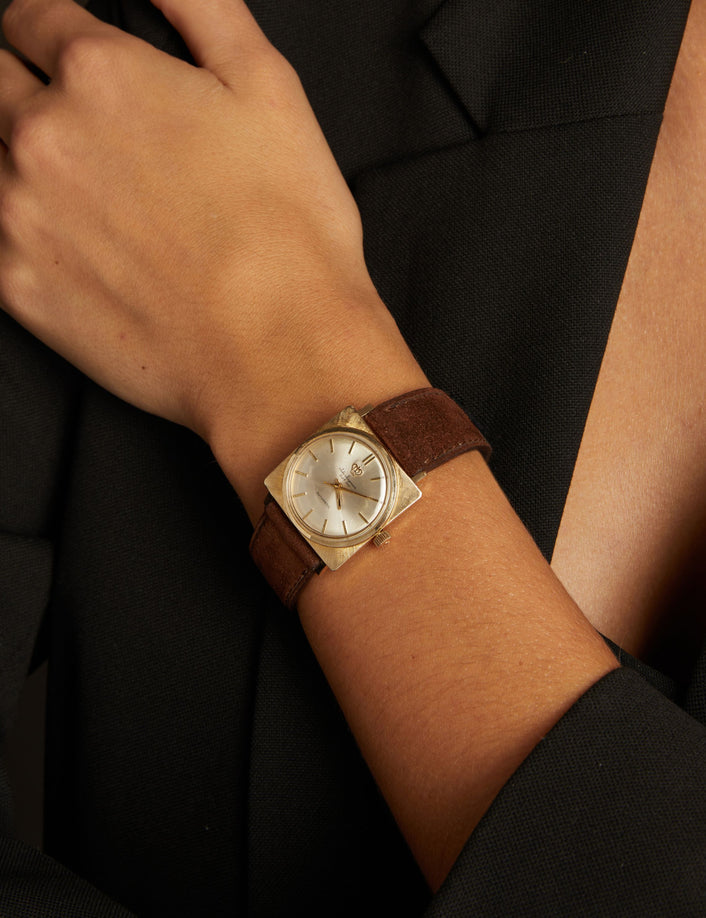 Model. Close up of a woman wearing a brown leather watch with gold face on neutral background.
