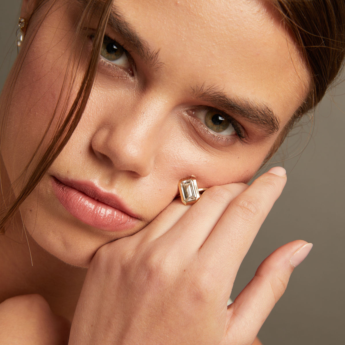 Model. Close-up of a woman with an oval diamond bezel ring resting on her cheek and hand, against a neutral gray background.