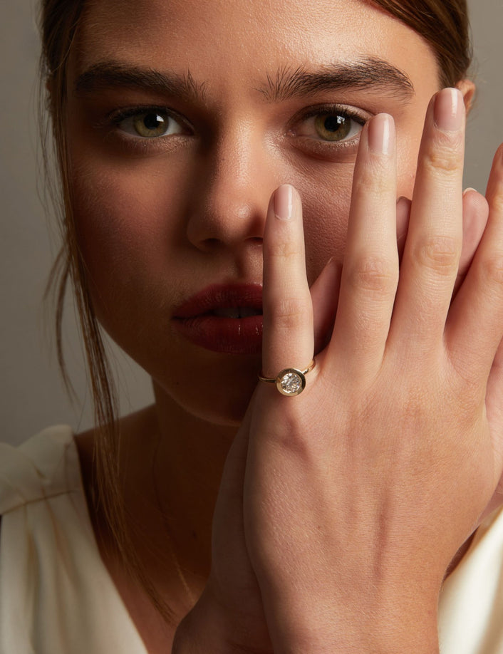 Model. A close-up lifestyle shot of a woman wearing a delicate gold ring with a small round clear gemstone on her finger, posed against a soft neutral background.