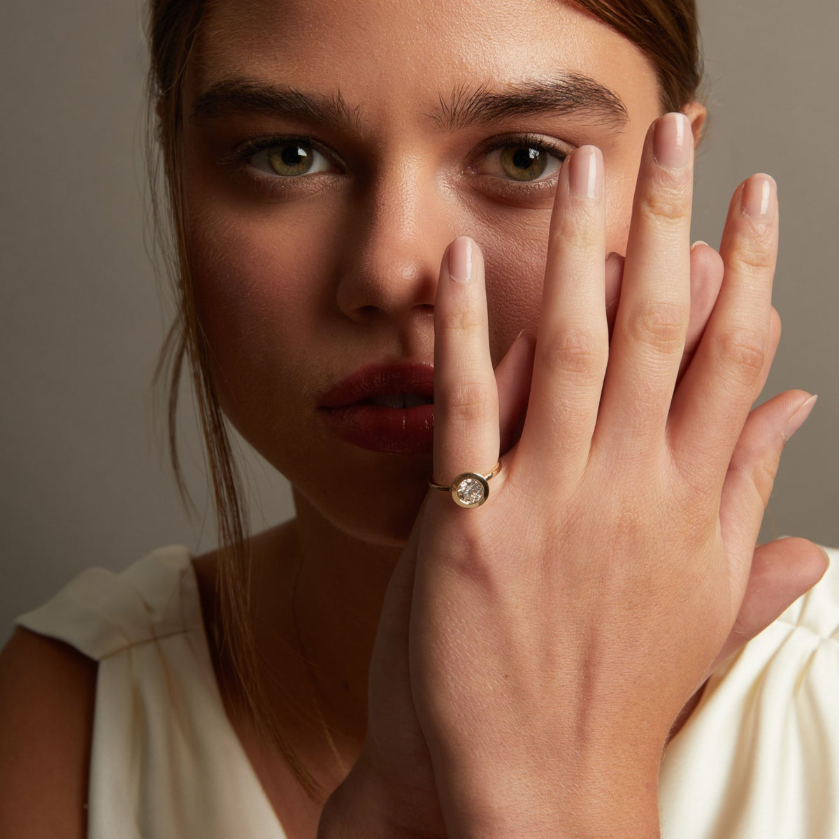 Model. A close-up lifestyle shot of a woman wearing a delicate gold ring with a small round clear gemstone on her finger, posed against a soft neutral background.