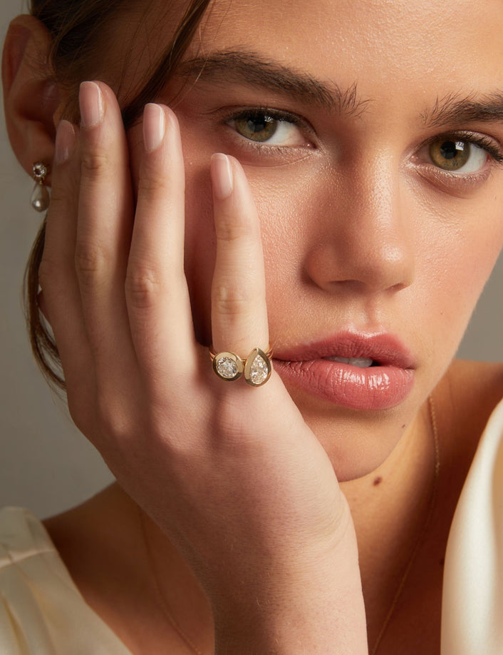 Model. A close-up lifestyle shot of a model wearing two gold rings with a pear and round diamond, with matching pearl drop earrings visible on her ear lobe.