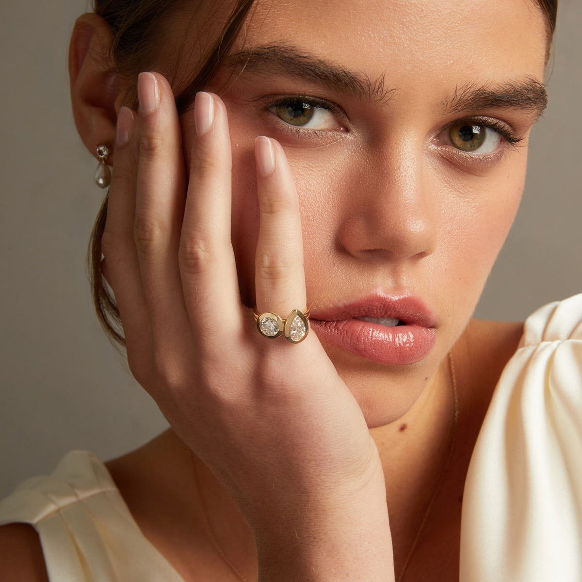 Model. A close-up lifestyle shot of a model wearing two gold rings with a pear and round diamond, with matching pearl drop earrings visible on her ear lobe.