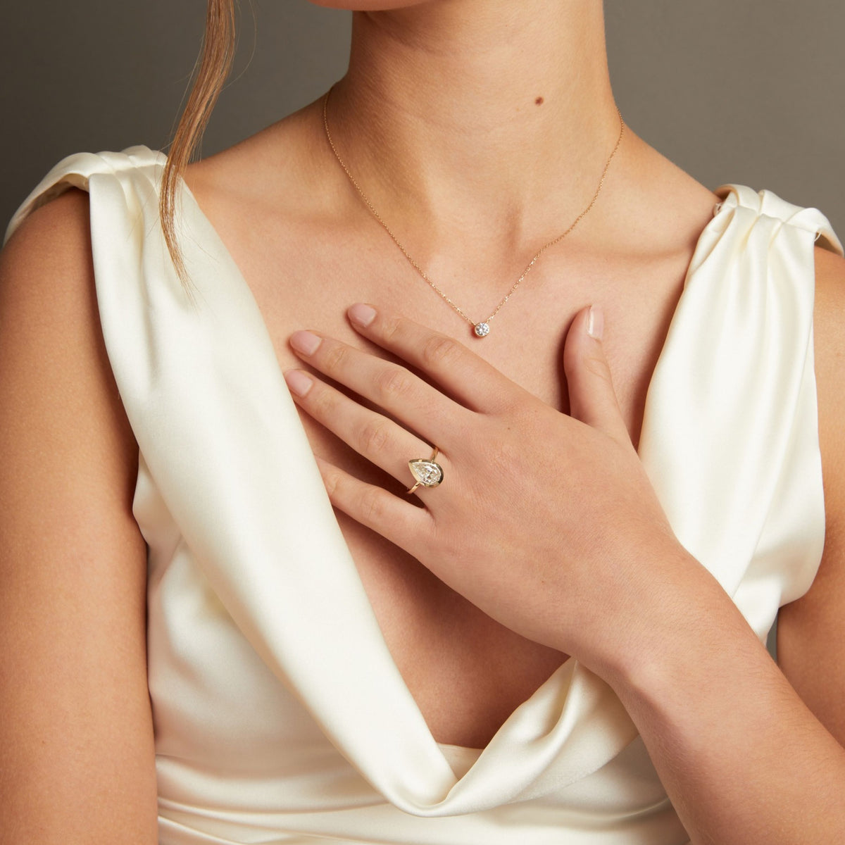 Model. A close-up lifestyle shot of a woman's hand wearing a gold ring with a pear bezel-set diamond. She is wearing a cream satin top against a neutral gray background.
