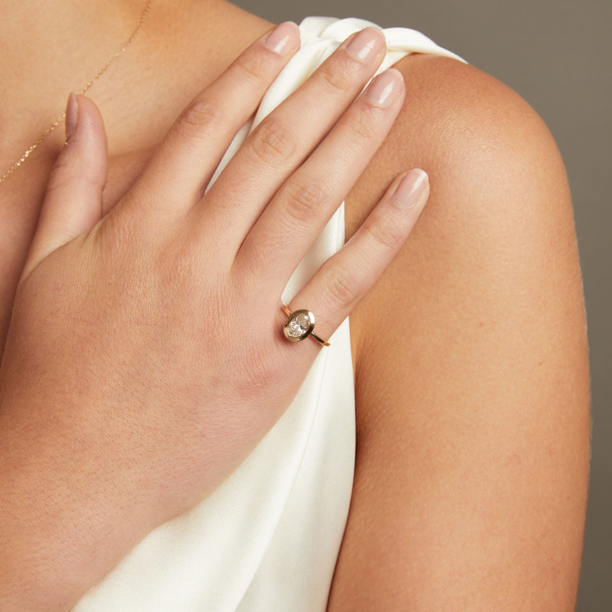 Model. A close-up lifestyle shot of a woman's hand resting on her shoulder, wearing a delicate gold ring with a small clear oval shaped diamond on a gold band.