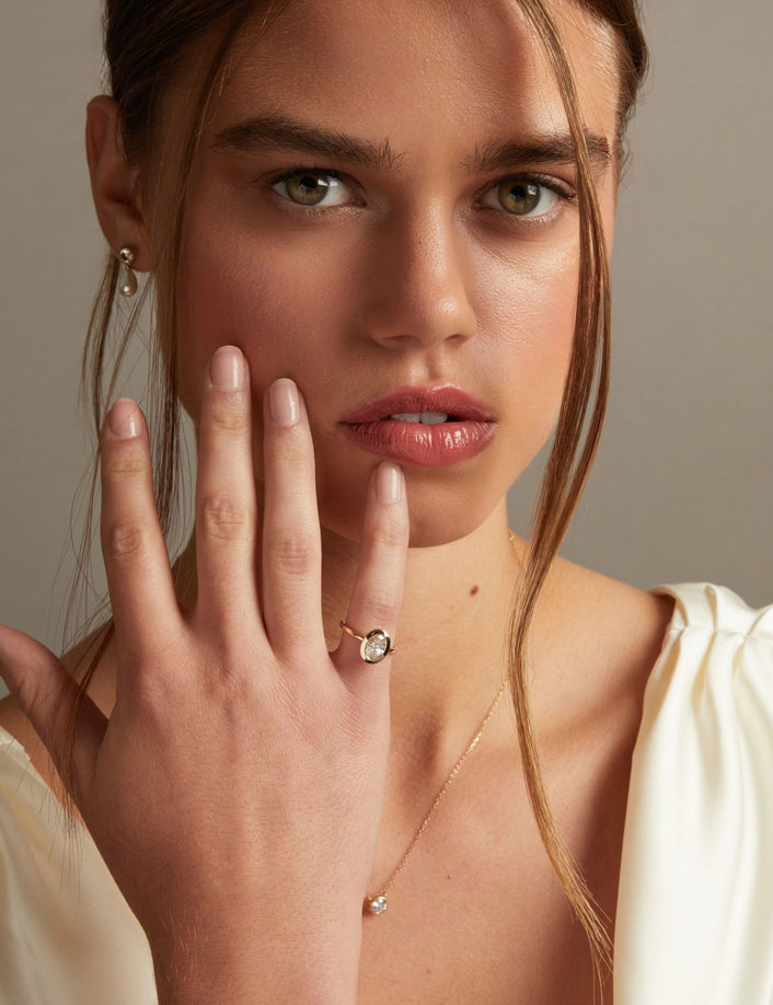 Model. A close-up lifestyle shot of a woman wearing a delicate gold ring with an oval clear diamond on her finger, paired with a matching gold necklace. The background is a soft, neutral gray.