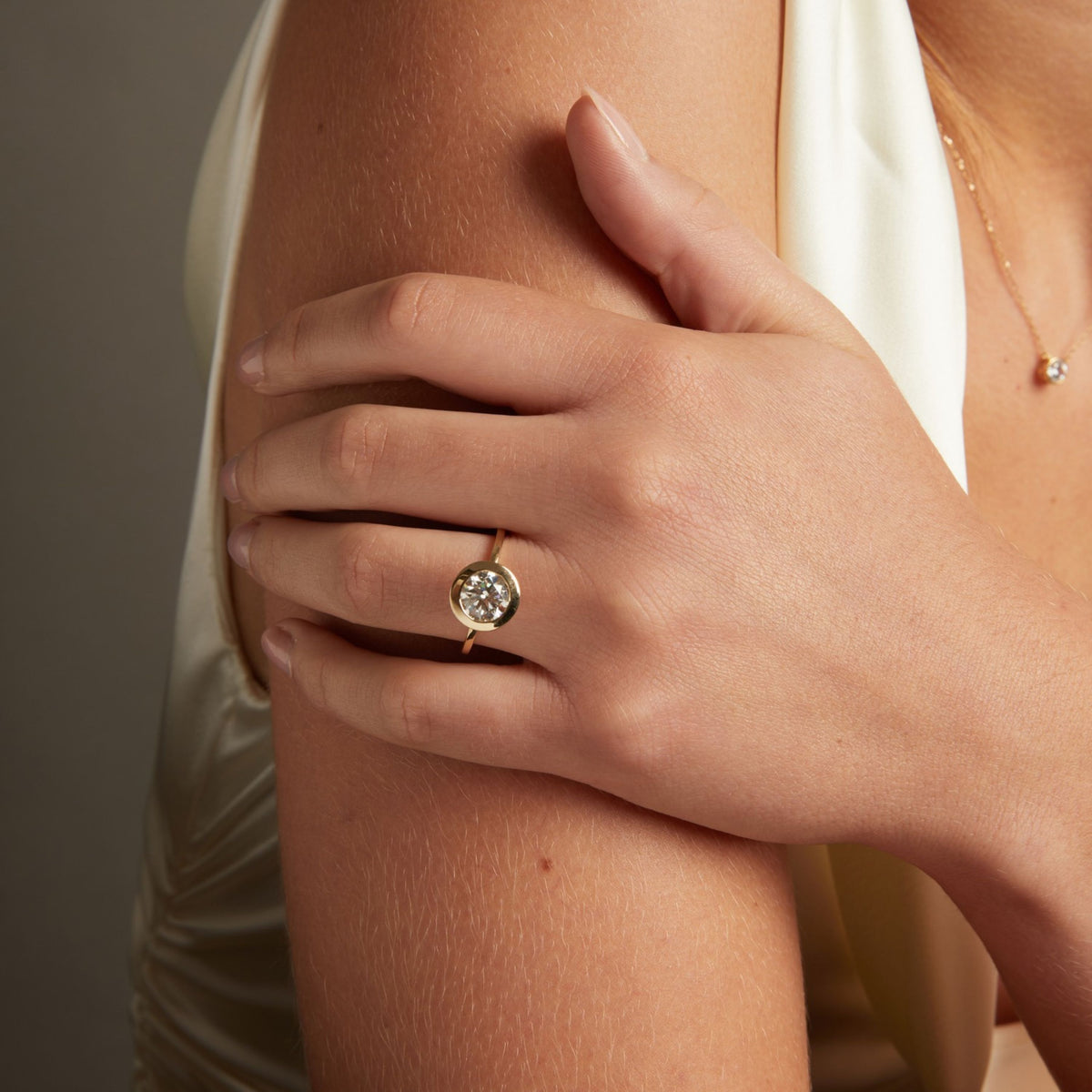 Model. A close-up lifestyle shot of a woman's hand resting on her shoulder, wearing a gold ring with a round bezel-set diamond. She is wearing a cream satin top against a neutral gray background.