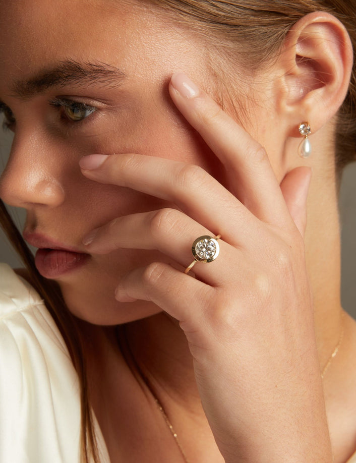 A close-up lifestyle shot of a woman wearing a round bezel set diamond ring, posed against a soft neutral background.