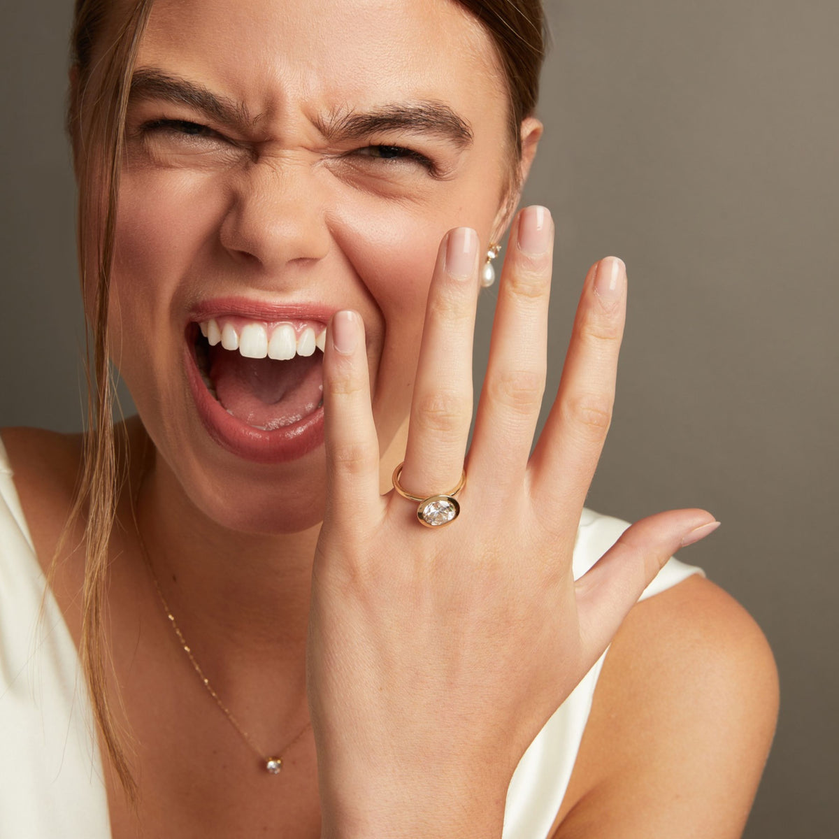 Model. A close-up lifestyle shot of a woman smiling and showing her hand with a round bezel set diamond ring, posed against a soft neutral background.