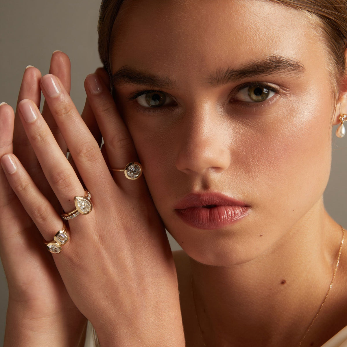 Model. A close-up lifestyle shot of a woman wearing various diamond bezel rings, posed against a soft neutral background.