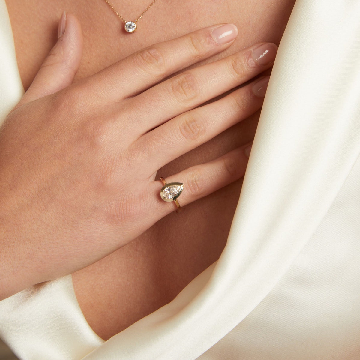 Model. A close-up lifestyle shot of a woman's hand resting on her chest, wearing a gold ring with a pear shaped diamond. She is also wearing a delicate gold necklace with a single round diamond, set against a soft cream-colored satin fabric.