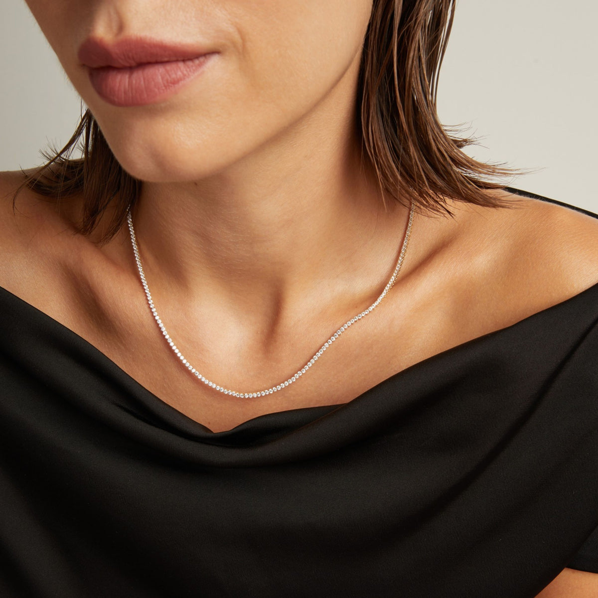 Close up of a woman in a black top wearing a diamond tennis necklace on beige background.