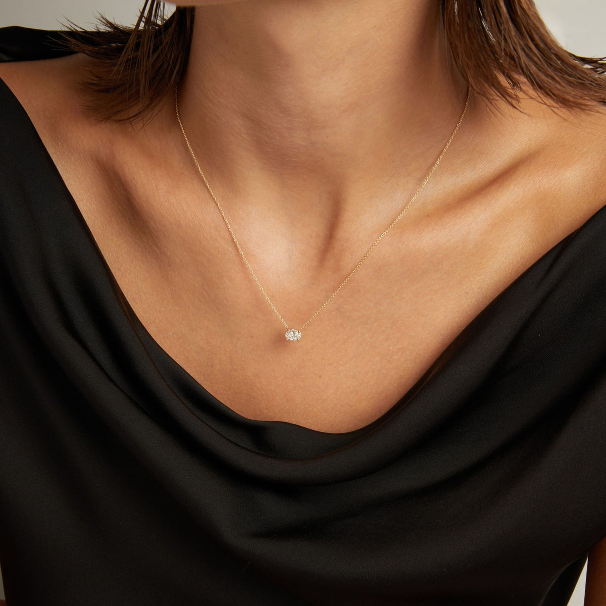 Model. Close up of woman wearing a black top with an oval diamond solitaire necklaces on a beige background.