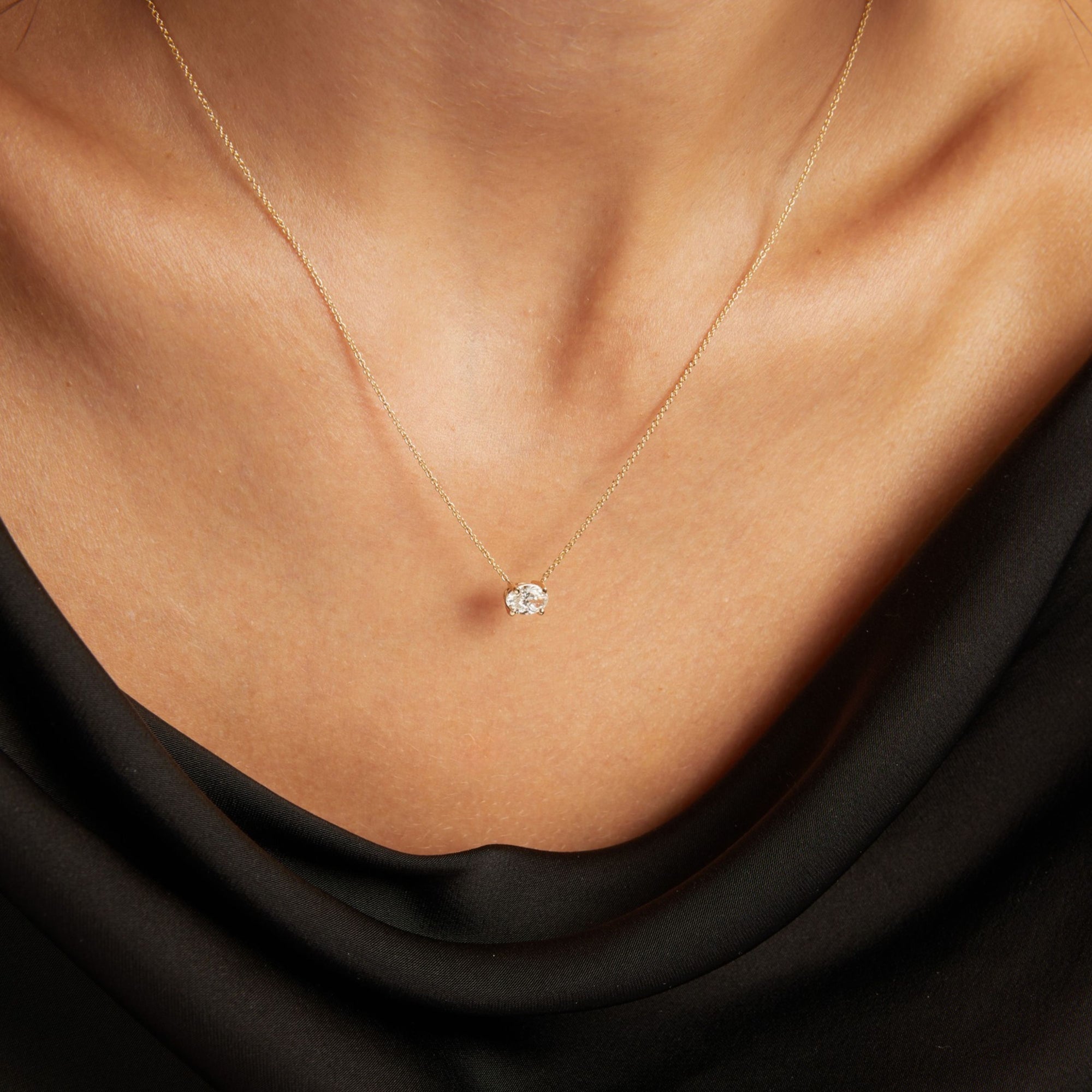 Close up of woman wearing a black top with an oval diamond solitaire necklaces on a beige background.