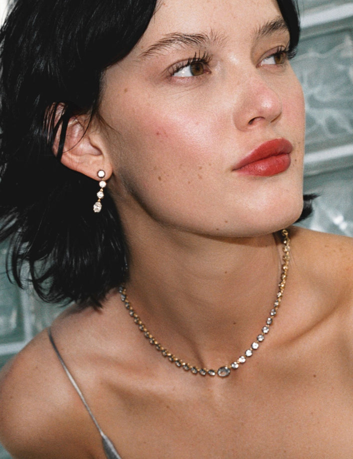 Model. Close up of woman in silver dress wearing a gemstone tennis necklace and matching earrings on against a glass window. 