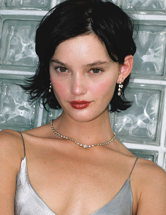 Model. Close up of woman in silver dress wearing a gemstone tennis necklace and matching earrings on against a glass window. 