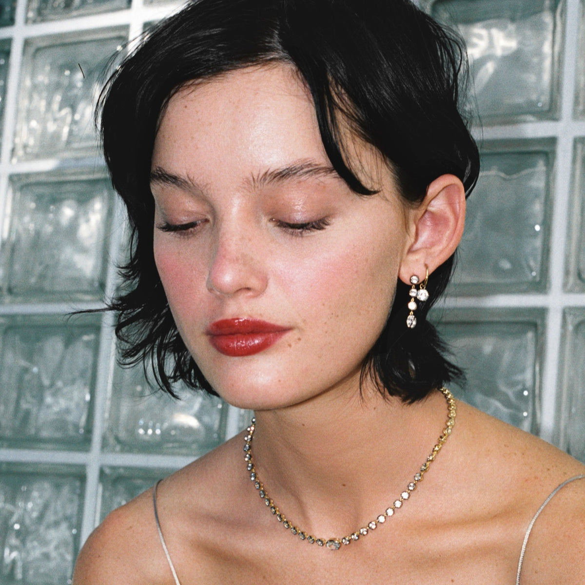 Model. Close up of woman in silver dress wearing a gemstone tennis necklace and matching earrings on against a glass window. 