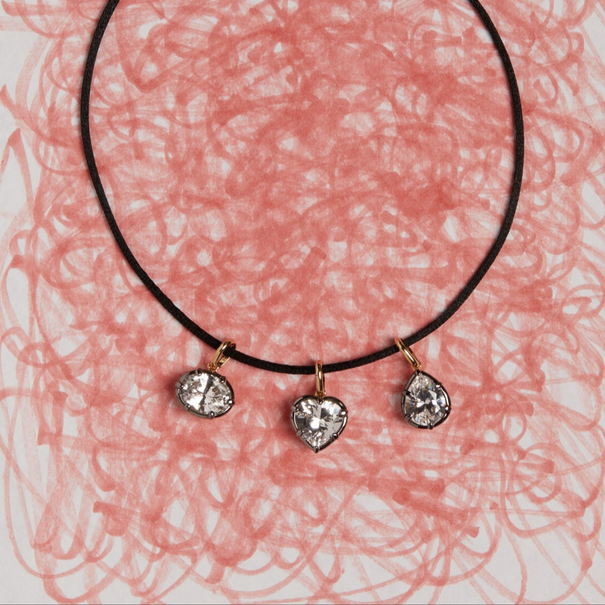 Model. Close up of cord necklace with three gemstone pendants laying on white sheet of paper with pink scribbles. 