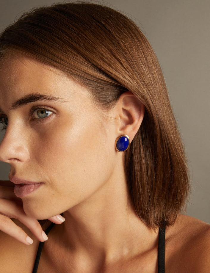 Close up of a woman wearing a large blue gemstone stud earring with gold accents on neutral background.