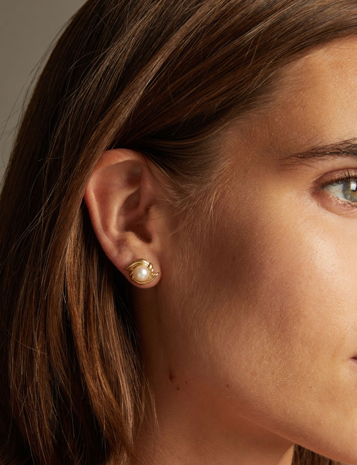 Close up of a woman wearing a pearl stud earring with gold accents on neutral background.
