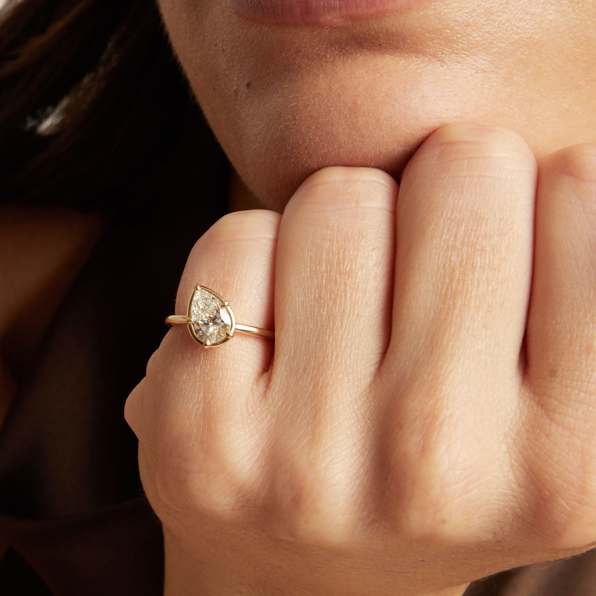 A close-up lifestyle shot of a woman wearing a delicate gold ring with a pear diamond. 