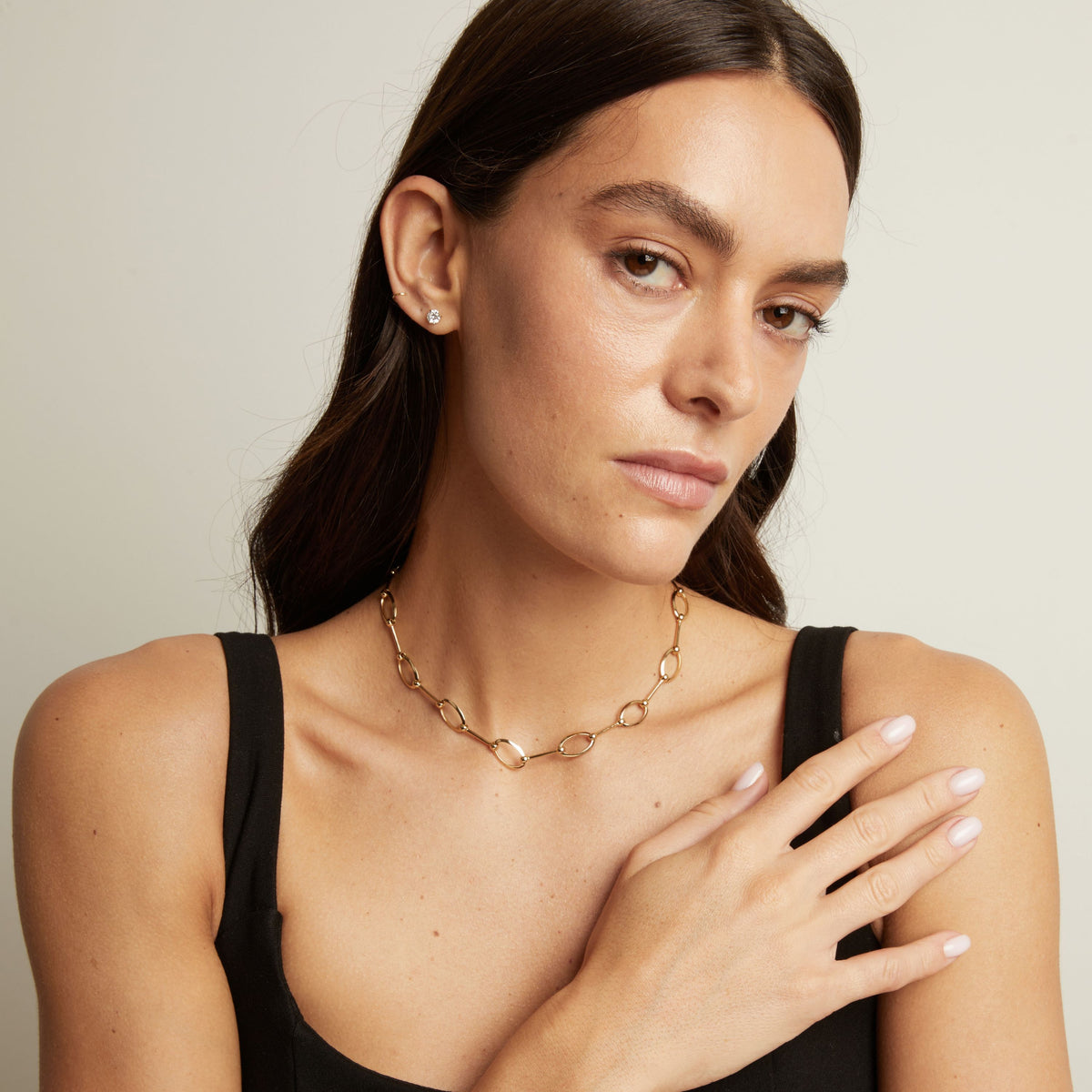 A close-up of a woman wearing a gold chain necklace with interlocking oval links, paired with a black tank top against a light gray background.