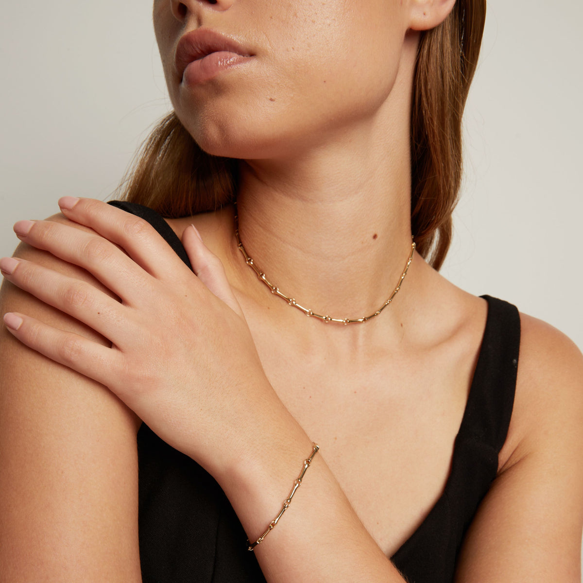 Model. A close-up of a woman wearing a delicate gold chain necklace and a matching gold bracelet, paired with a black top against a neutral light gray background.