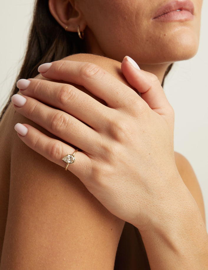 A close-up lifestyle shot of a woman wearing a delicate gold ring with a pear diamond. 