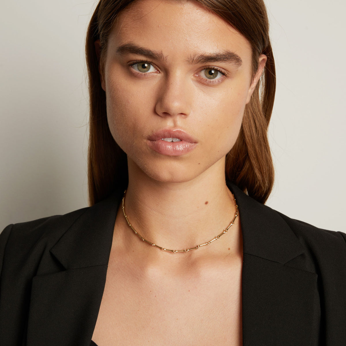 Model. A close-up of a woman wearing a delicate gold chain necklace with small links, styled with a black blazer against a neutral light gray background.