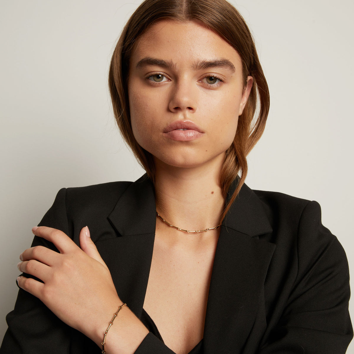 Model. A close-up of a woman wearing a delicate gold chain necklace with small links, styled with a black blazer against a neutral light gray background.