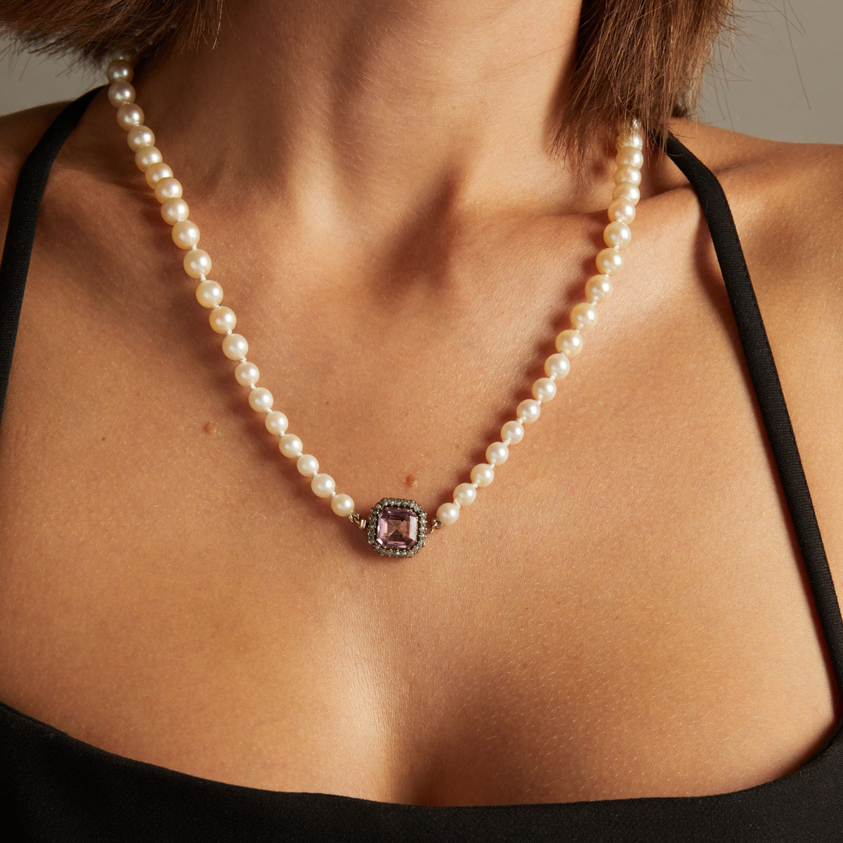 Model. Close up of a woman wearing a pearl necklace with a bold pink gemstone on neutral background.