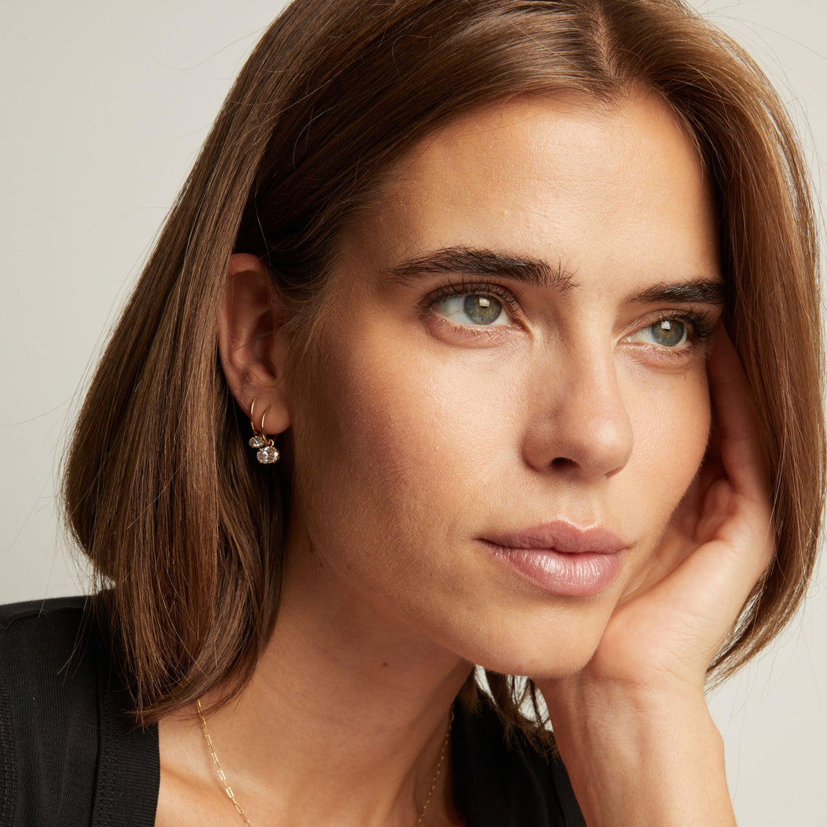 Model. Close up of a woman in a black top wearing two oval diamond huggie earrings on neutral background.