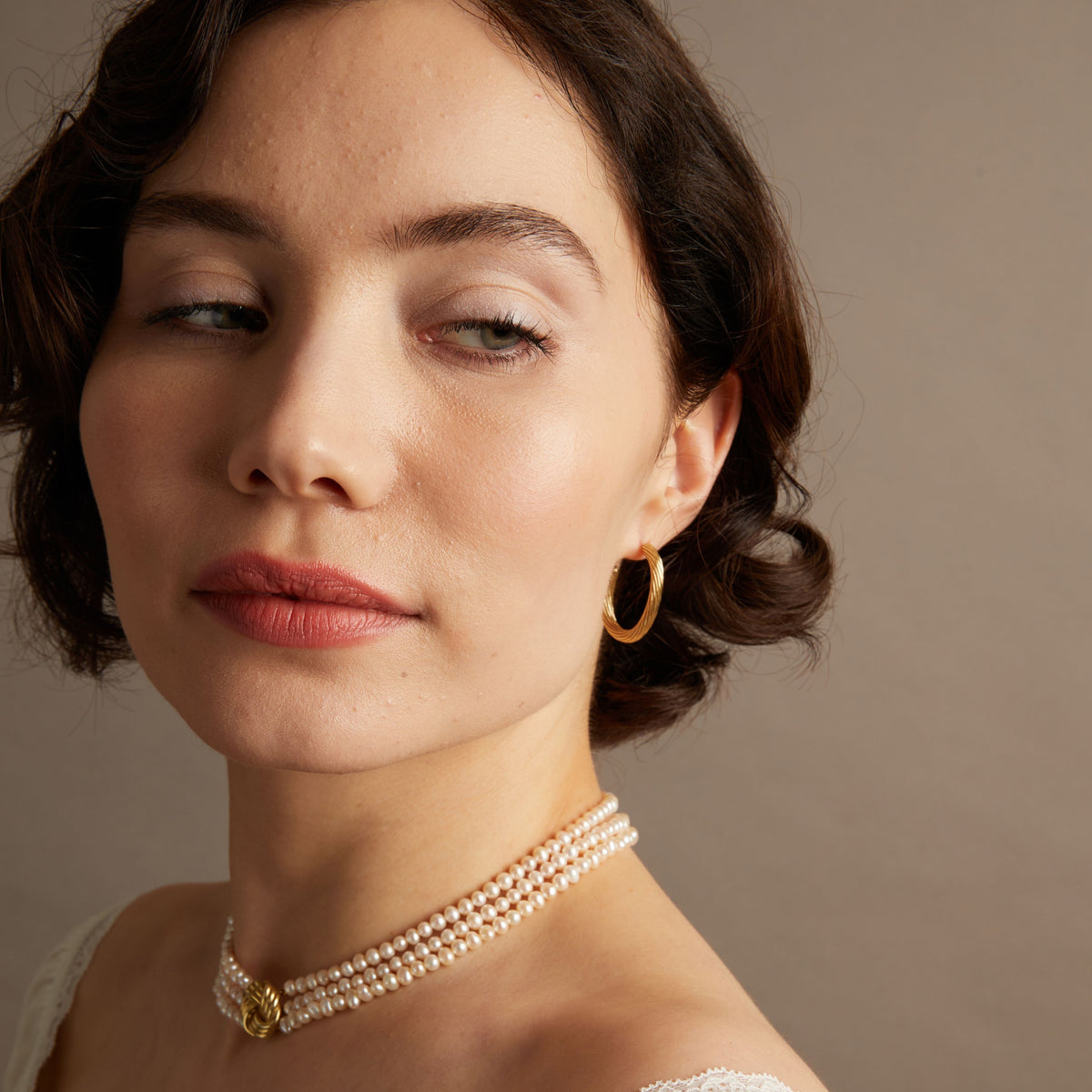 Woman wearing gold hoop earrings and pearl necklace with neutral background.