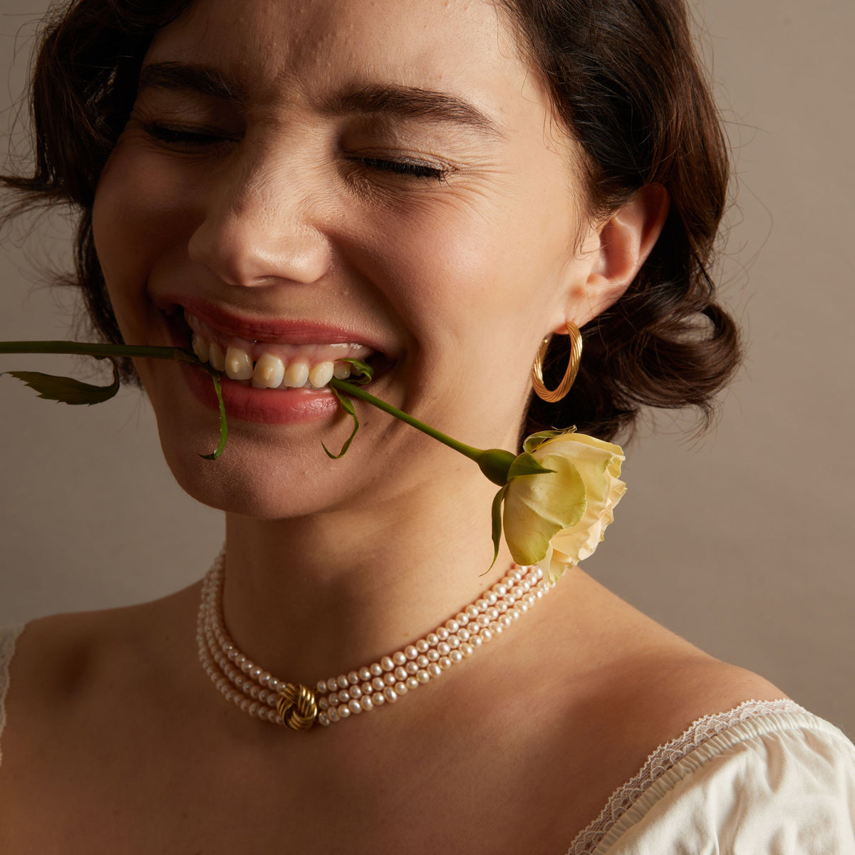 Woman wearing a pearl necklace and gold hoop earrings with a yellow rose stem in her mouth against a neutral background. 
