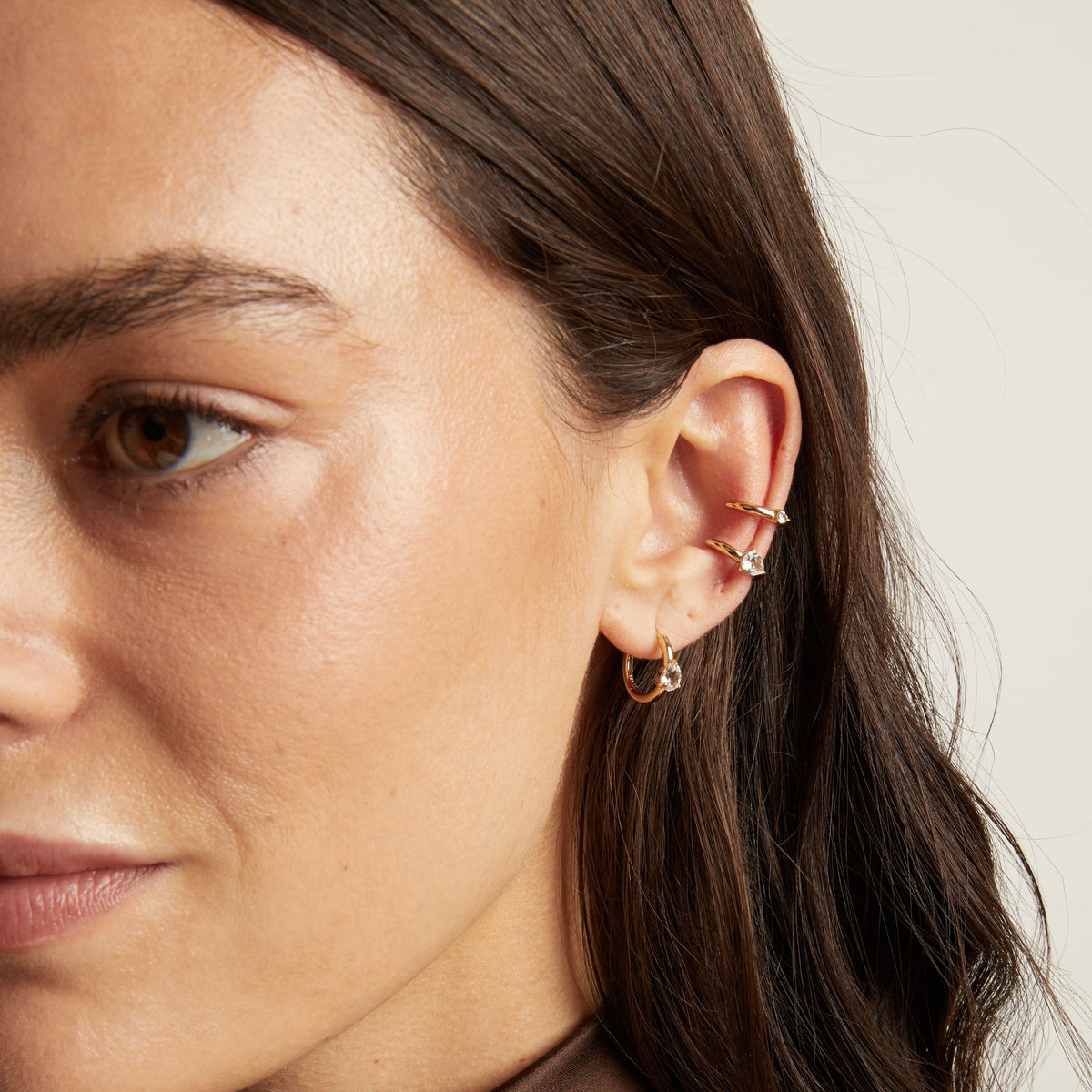 Close up of woman wearing two gold ear cuffs and a huggie earring with white sapphires on neutral background. 