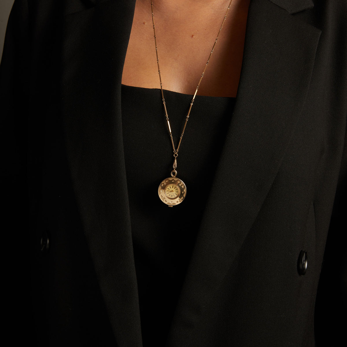 Close up of a woman wearing a large watch pendant necklace on neutral background.