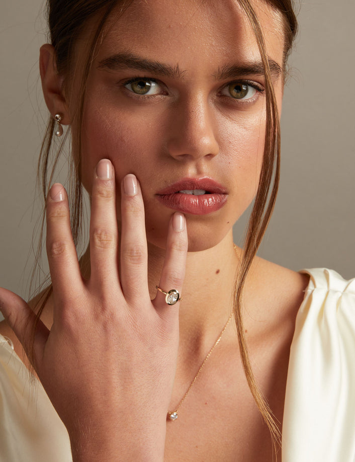 Model. A close-up lifestyle shot of a woman wearing a delicate gold ring with an oval clear diamond on her finger, paired with a matching gold necklace. The background is a soft, neutral gray.